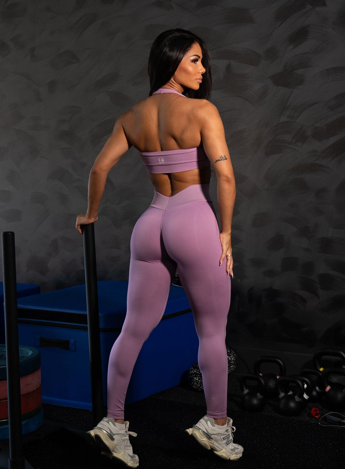 Woman wearing a mauve halter sports bra and matching high-waisted leggings, shown from behind next to a weight sled in a gym setting.