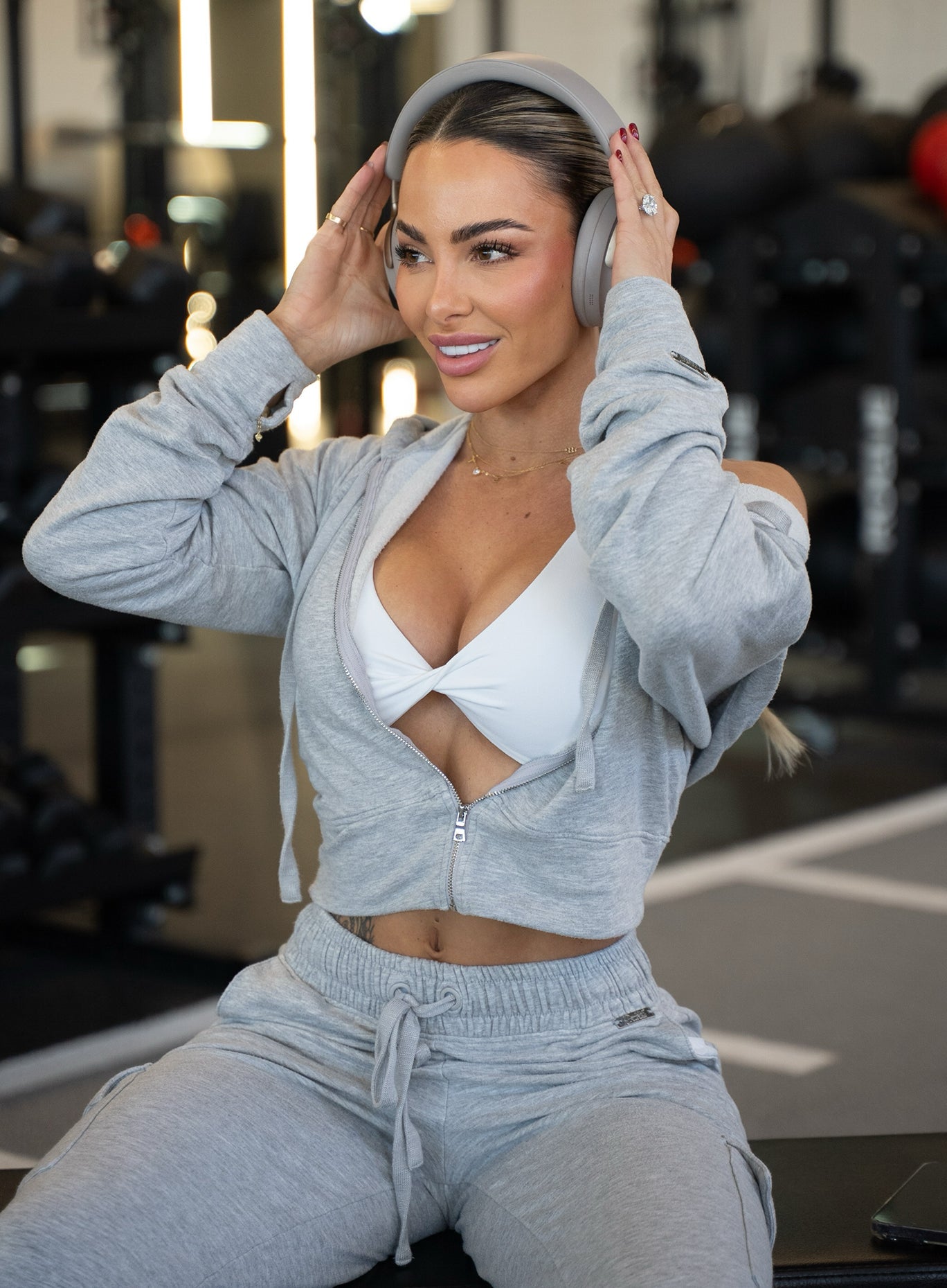Woman wearing a grey cropped zip-up hoodie over a white sports bra, matching grey joggers, and over-ear headphones in a gym setting.