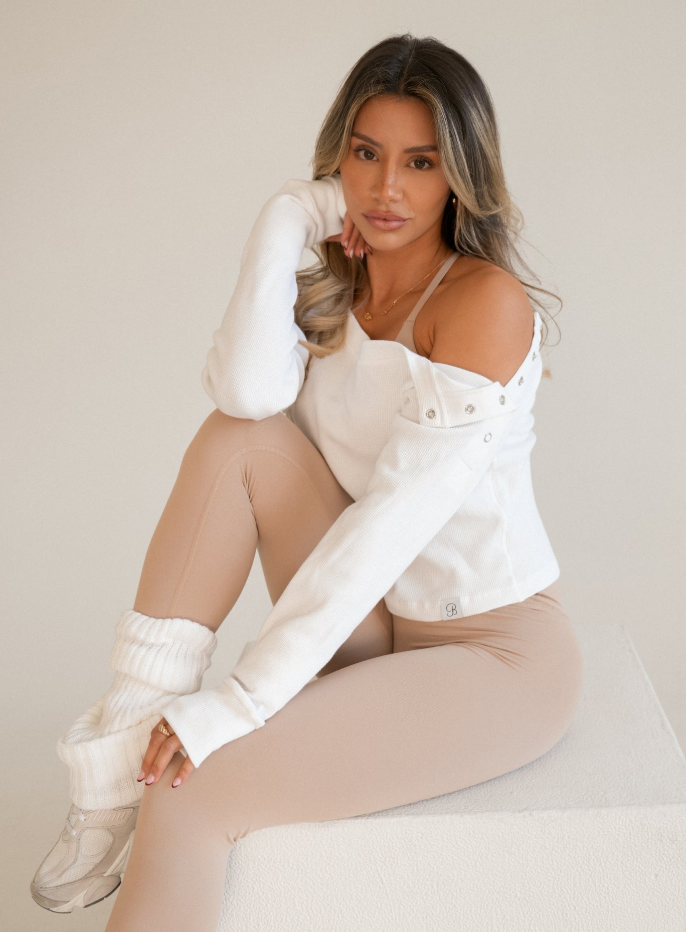 Woman wearing a white waffle-knit off-shoulder pullover with snap-button cuffs, paired with tan leggings and white knit leg warmers.
