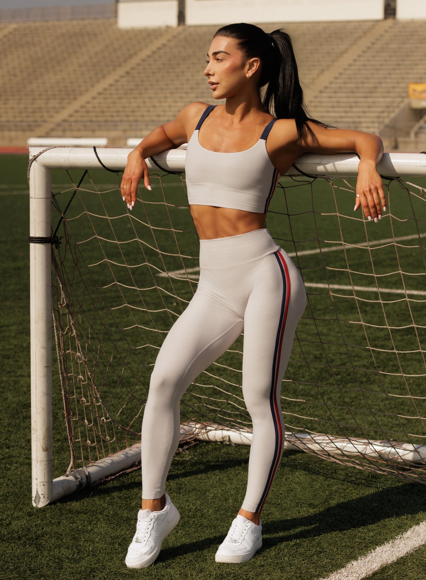 Woman wearing an off-white sports bra with navy straps and matching high-waisted leggings featuring a navy and red side stripe, standing at a soccer goal
