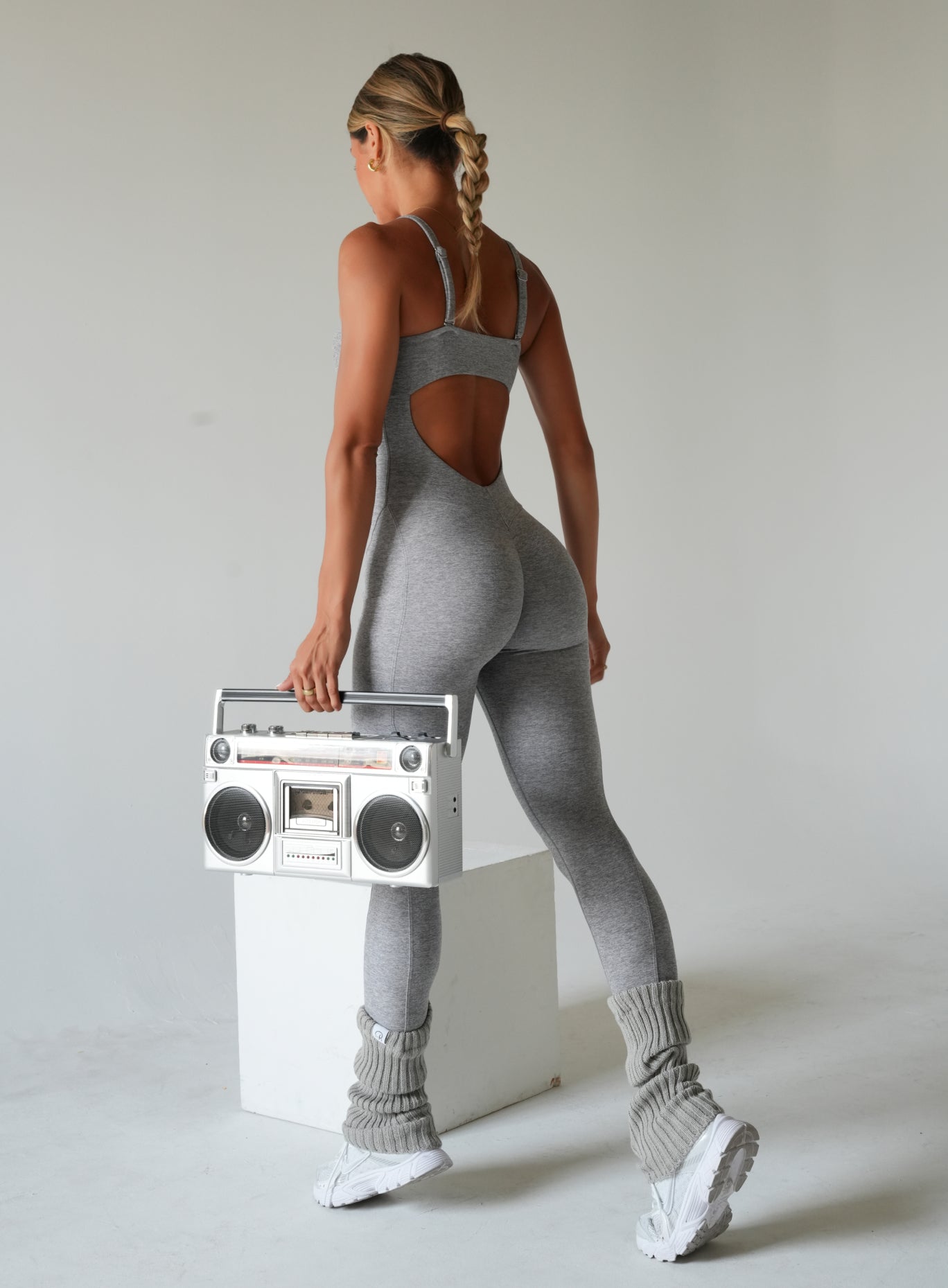 Back view of a woman wearing a heather gray fitted bodysuit with adjustable straps and a low open-back design, paired with gray leg warmers and white sneakers while holding a white retro boombox.