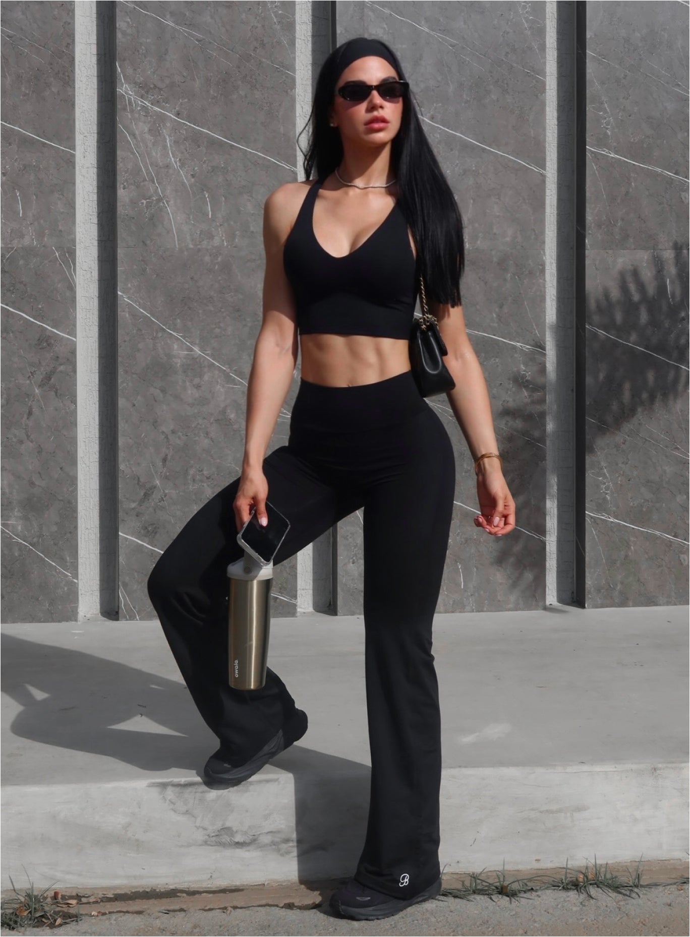 Woman wearing a black halter crop top and matching black flared pants, holding a water bottle and phone, standing against a marble tile wall.