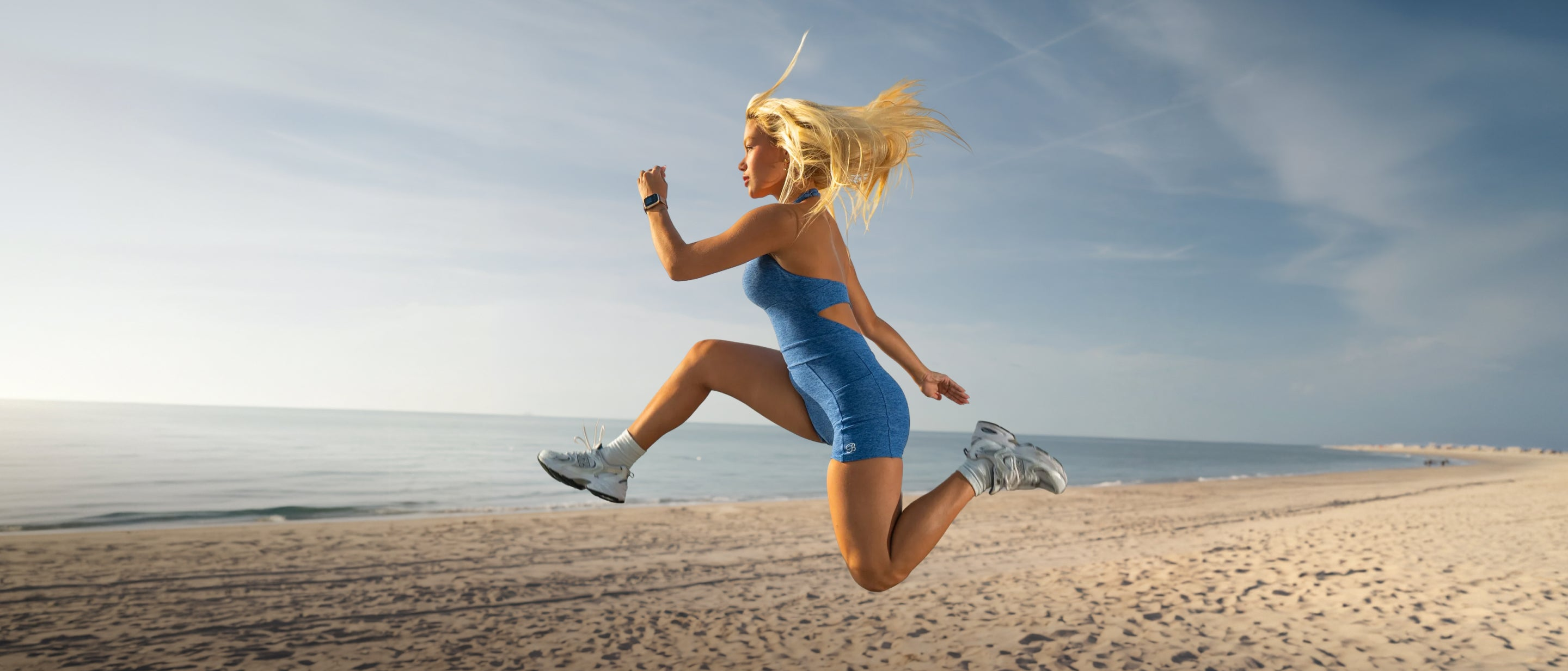 A woman in a blue strapless athletic dress jumps over the sand at a beach, with the ocean and blue sky in the background.