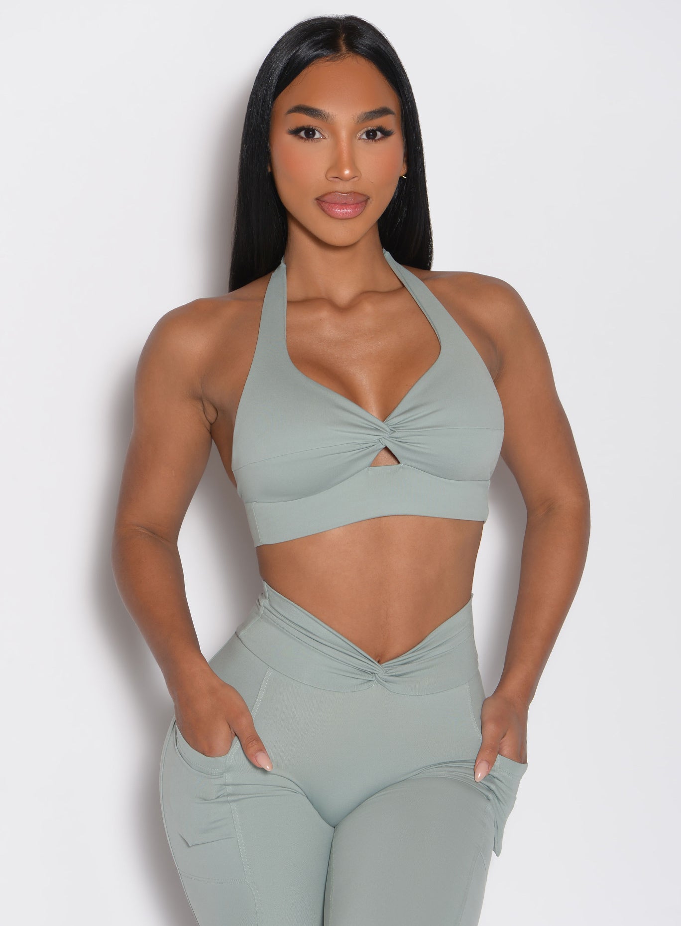 front profile view of a model with both hands inside the side pockets of the matching leggings wearing the Backless Knot Bra in Iceberg green color