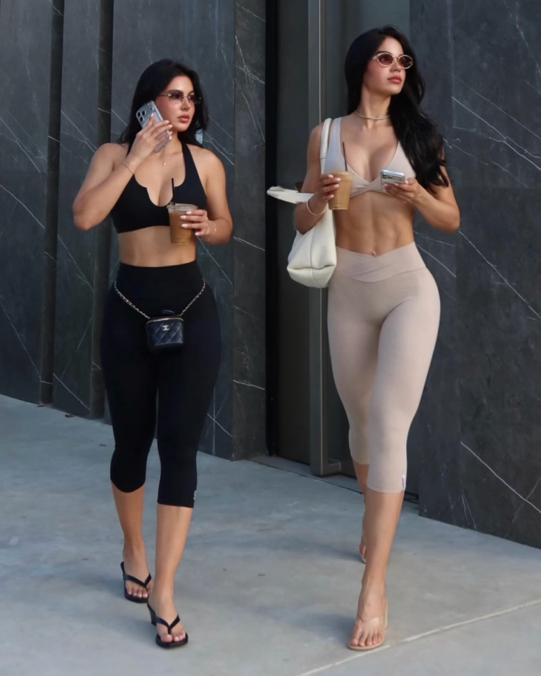 Two women walking outdoors in athletic wear. The woman on the left wears a black sports bra and black capri leggings with a small black crossbody bag.