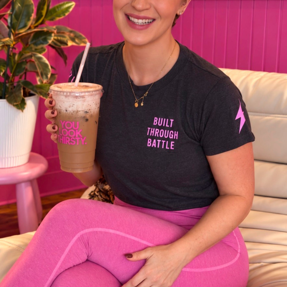 A woman sitting on a white couch wearing a dark charcoal gray t-shirt with pink text that reads 'BUILT THROUGH BATTLE' and a pink lightning bolt on the sleeve, paired with bright pink leggings. She is holding an iced coffee in a pink chair next to a potted plant.