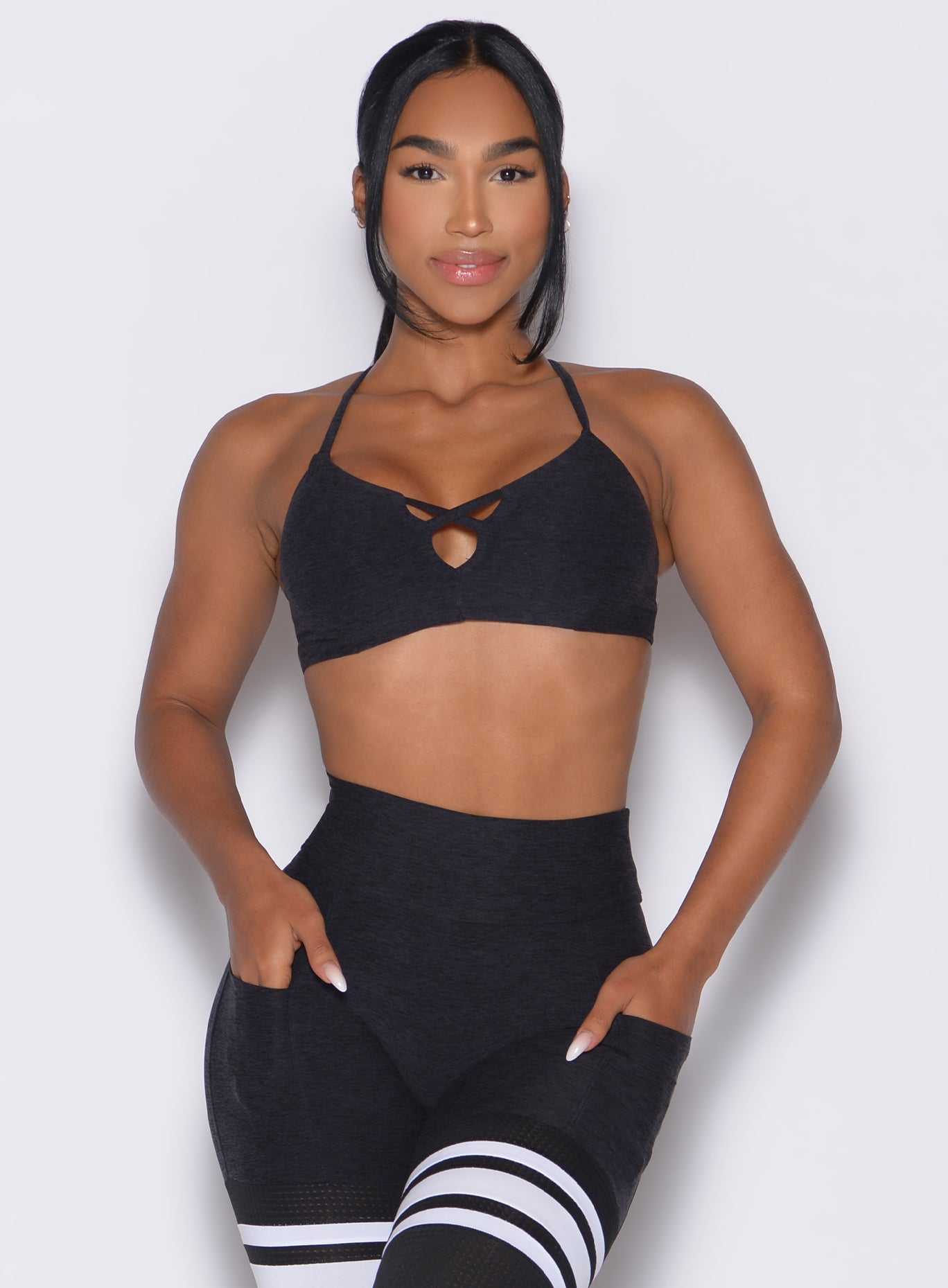 front profile view of a model with both hands inside the pockets of the leggings wearing the Lift Luxe Sports bra in Heathered black color