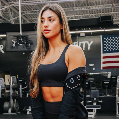 A woman in a gym wearing a dark gray sports bra and black athletic jacket, with a black sports bra and matching leggings visible. The background shows gym equipment and an American flag on the wall.