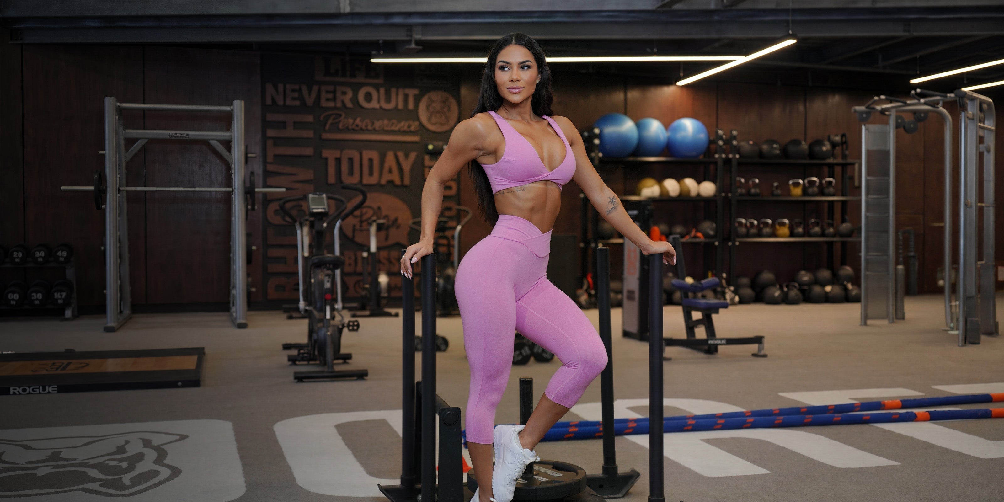 Woman posing in front of weights in a gym wearing a pink bra and matching capri leggings