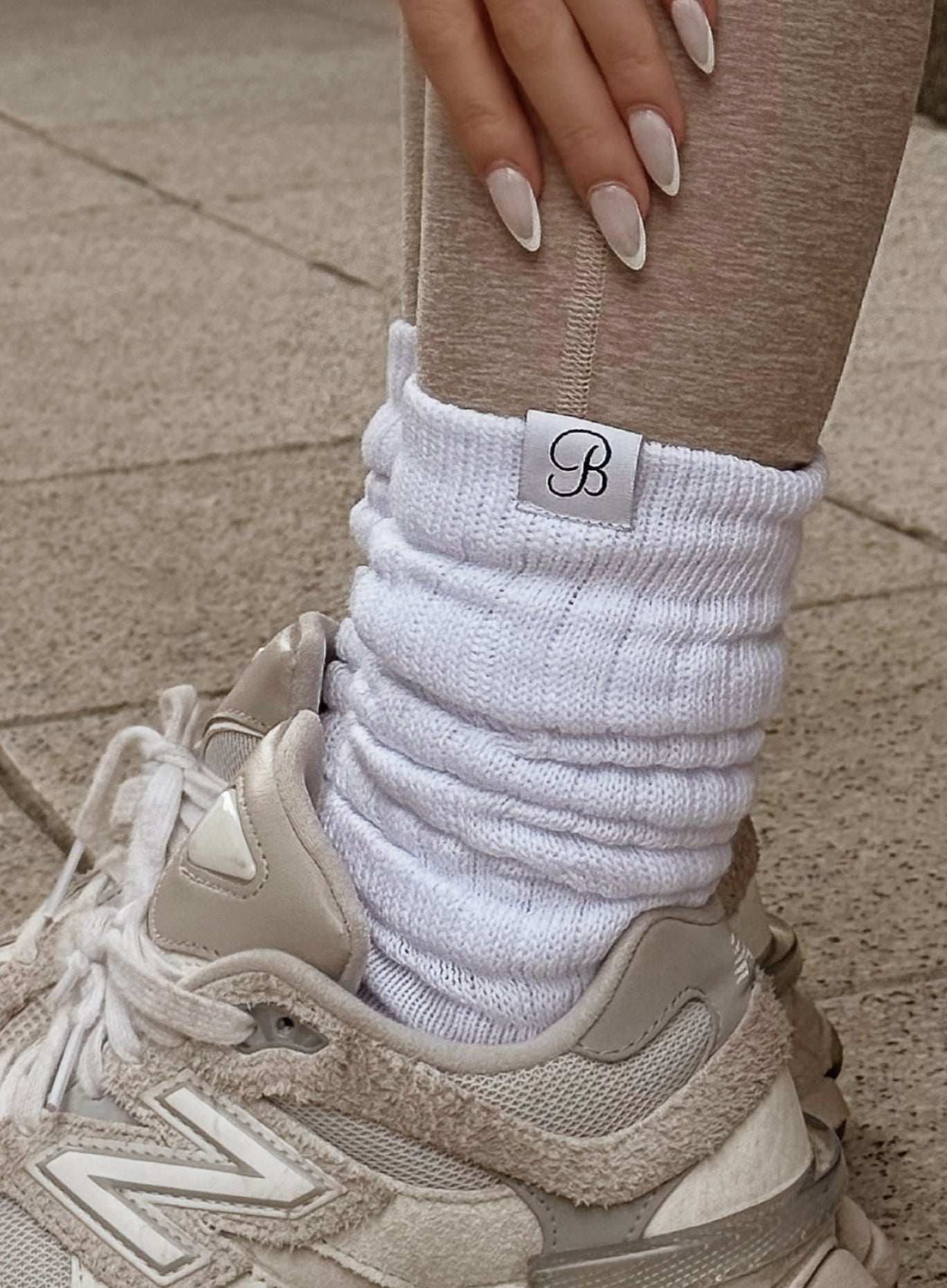 A close-up of a person's lower leg and foot wearing a white ribbed knit sock with a small white brand tab, paired with a beige sneaker and beige leggings.