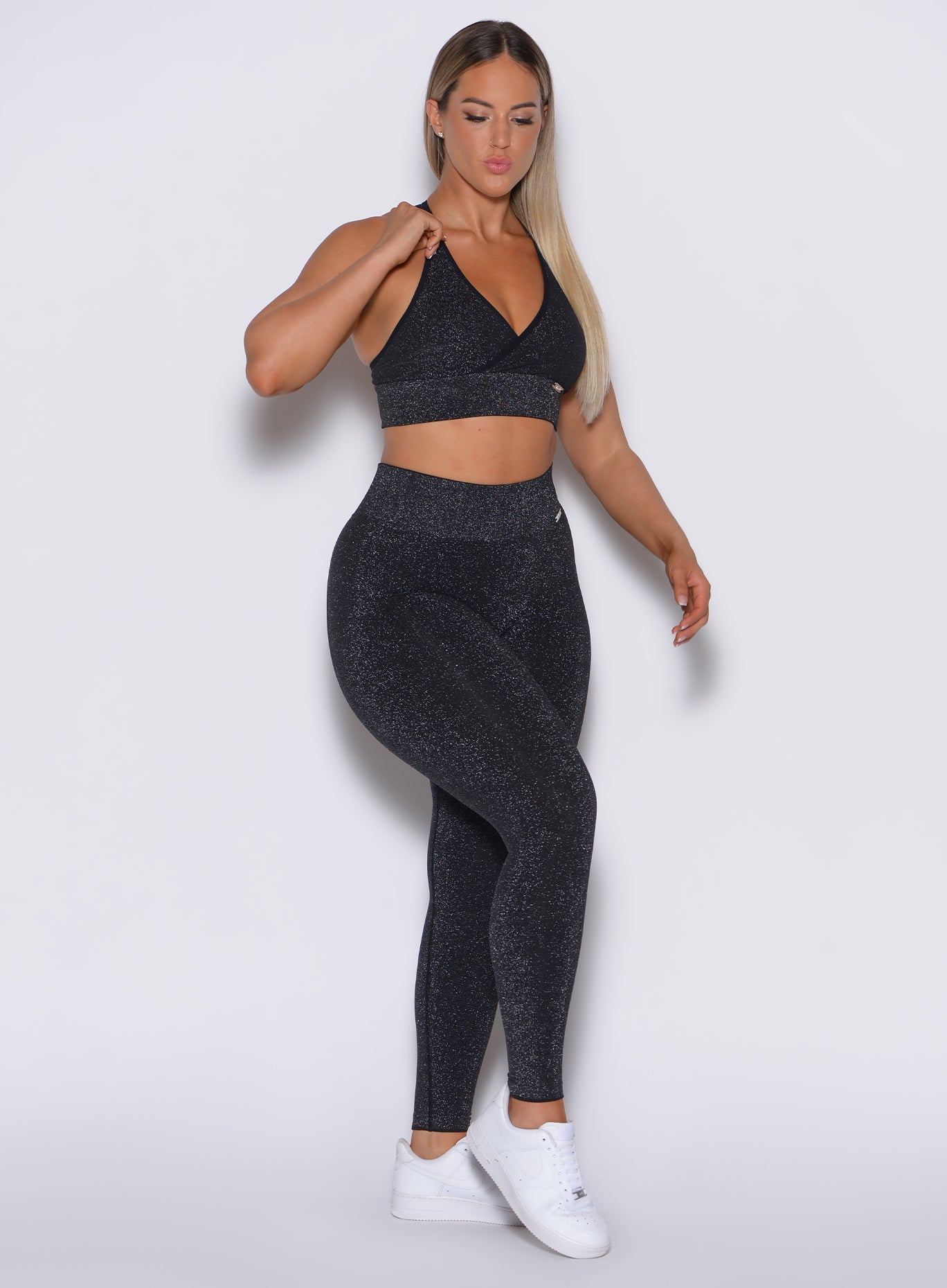 front side profile view of a model holding her left bra strap wearing our Shimmer Seamless Leggings in black sparkle color along with the matching bra
