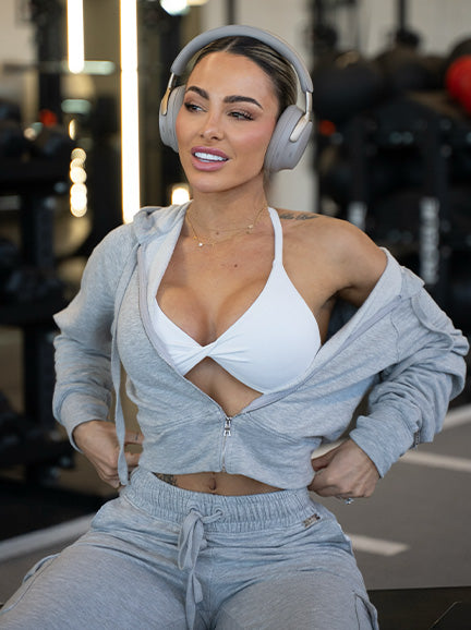Woman in a gym wearing a gray tracksuit and white sports bra, sitting on a bench with headphones on.
