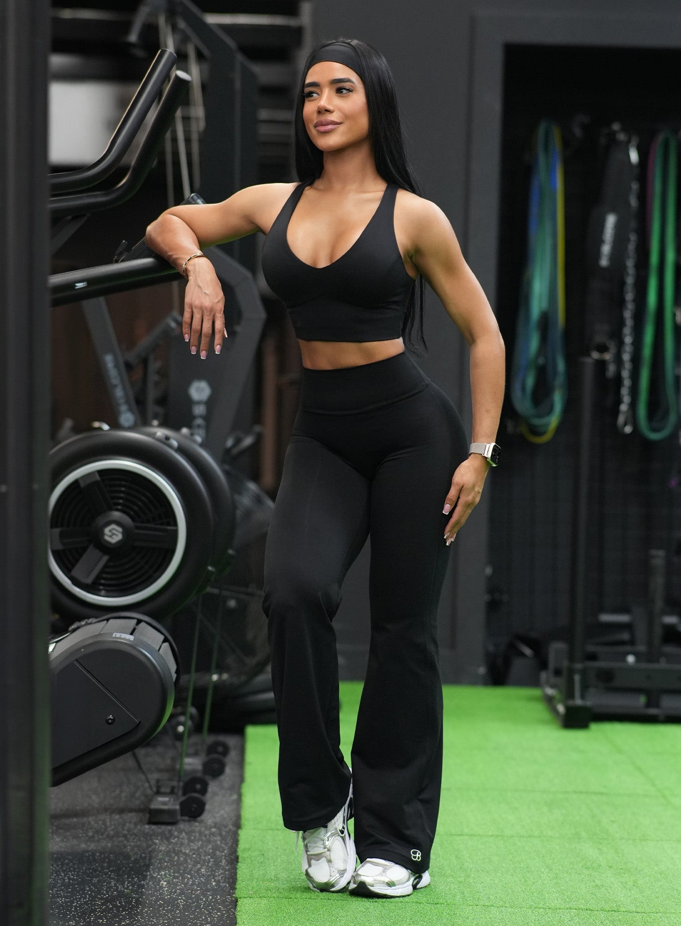 A woman posing in a gym wearing a matching black activewear set consisting of a scoop-neck sports bra and high-waisted flared leggings, paired with white sneakers.