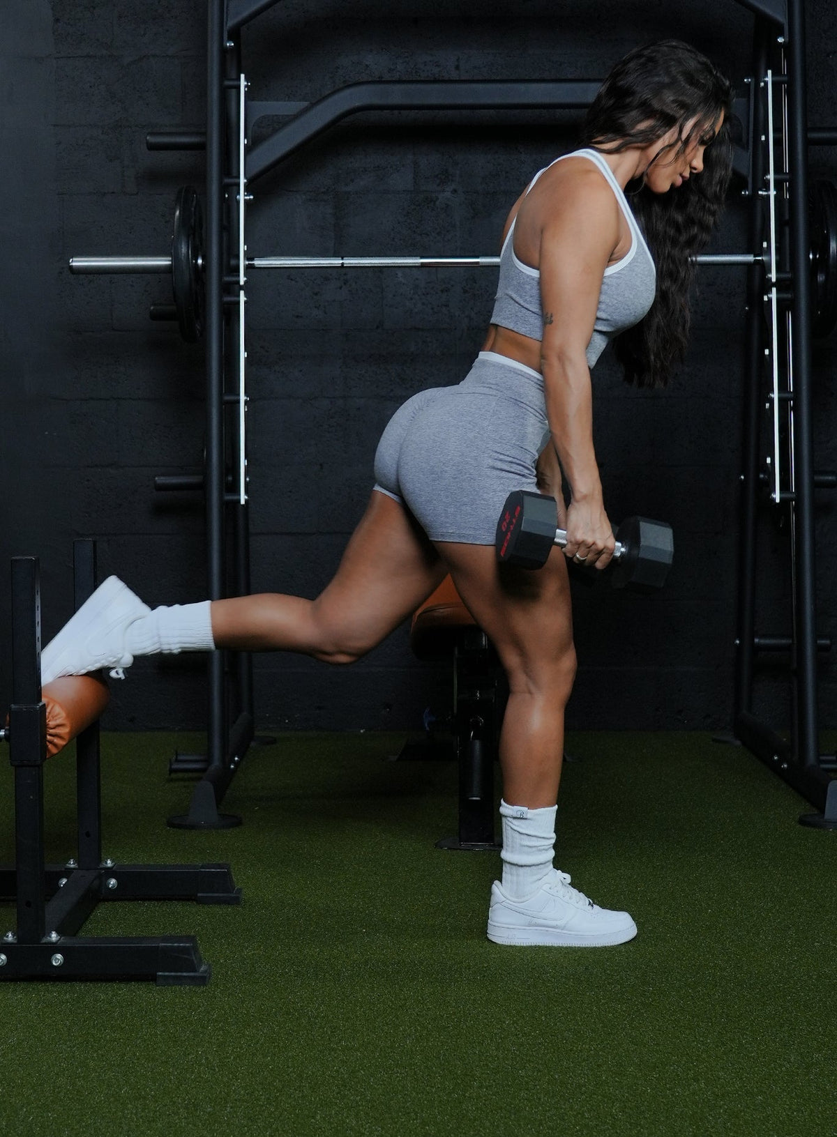Woman performing a Bulgarian split squat with a dumbbell, wearing a grey and white racerback tank and matching shorts.