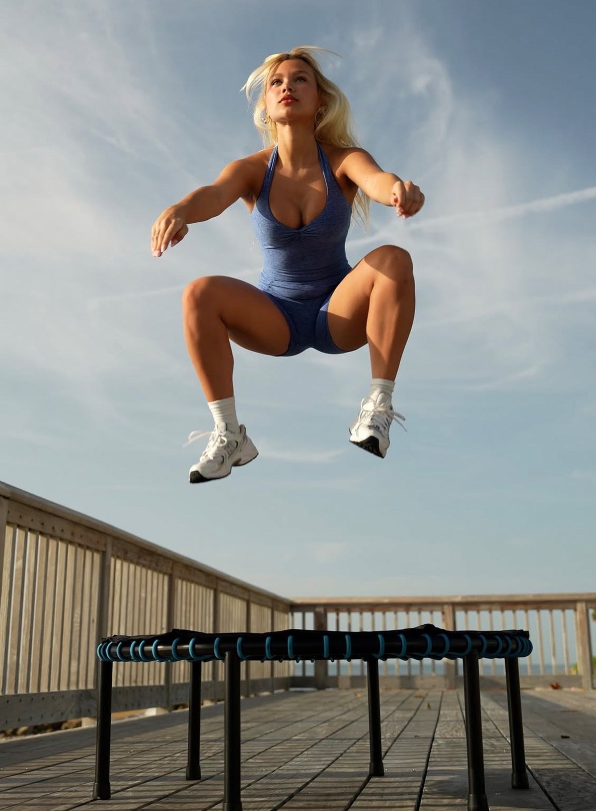 Woman jumping mid-air on a mini trampoline outdoors, wearing a blue halter bodysuit.