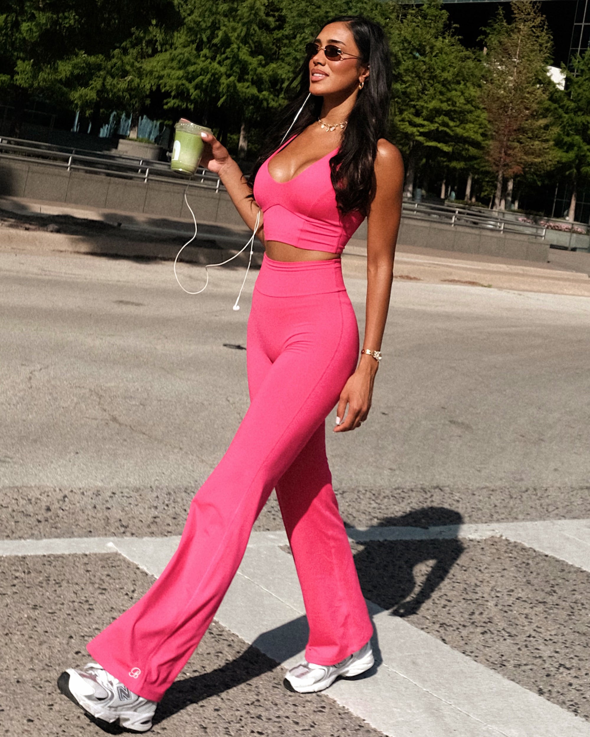 A woman walking outdoors on a crosswalk wearing a bright pink two-piece activewear set of a halter crop top and matching high-waisted flared pants, paired with white sneakers and holding a green drink.