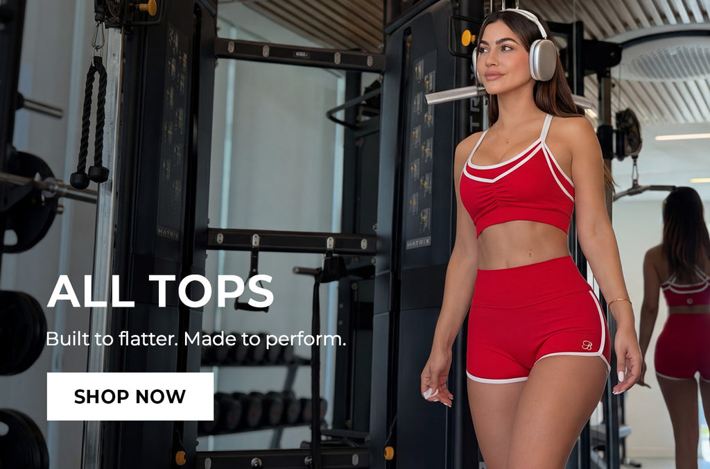 Woman in red athletic wear shorts and matching bra with headphones in a gym setting, promotional text overlay. Text says ALL TOPS Built to Flatter. Made to perform. SHOP NOW