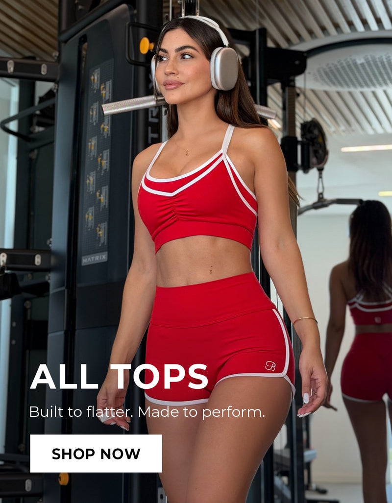 Woman in red athletic wear shorts and matching bra with headphones in a gym setting, promotional text overlay. Text says ALL TOPS Built to Flatter. Made to perform. SHOP NOW
