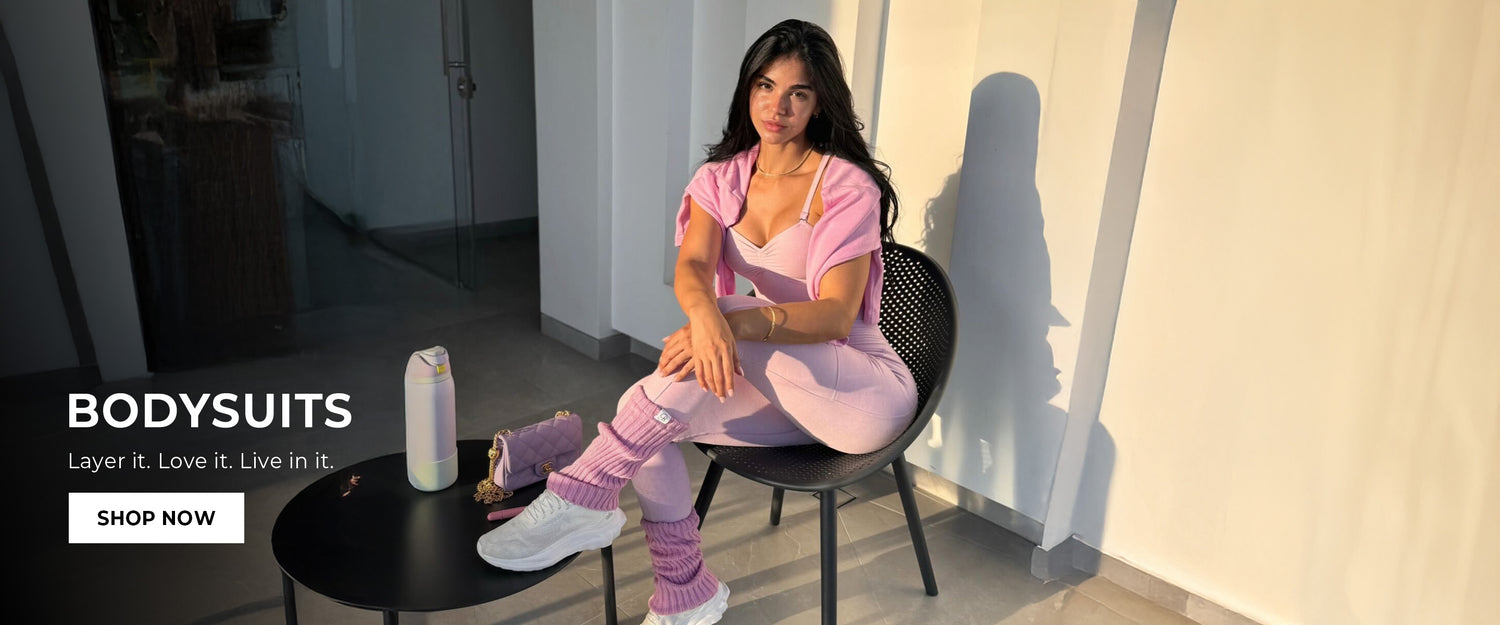 Woman in pink bodysuit sitting on a chair with promotional text for bodysuits. text reads: "BODY SUITS layer it. Love it. Live in it. SHOP NOW"