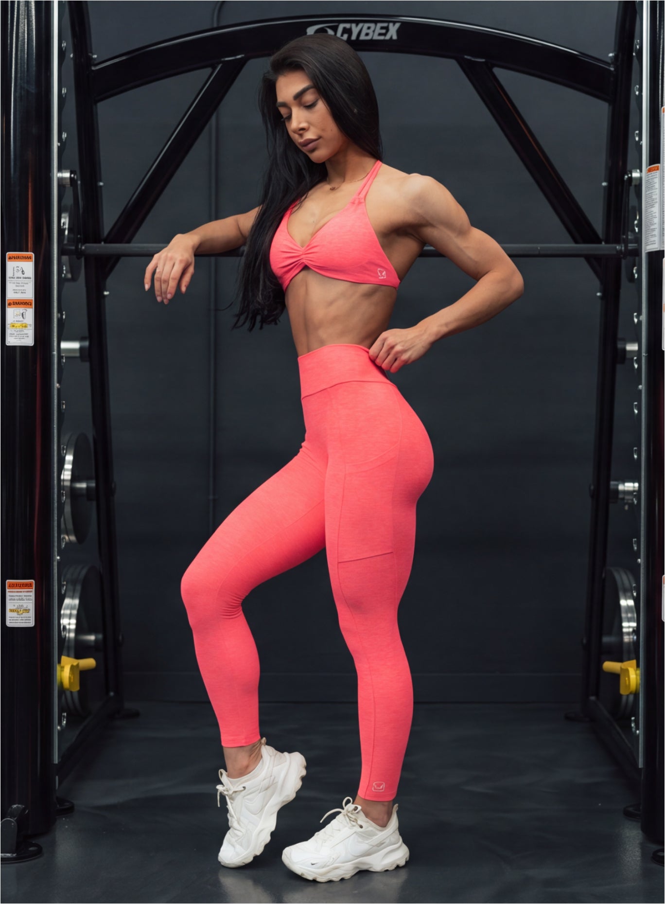 A woman posing in a gym wearing a matching coral pink activewear set consisting of a sports bra and high-waisted leggings, paired with white sneakers. 