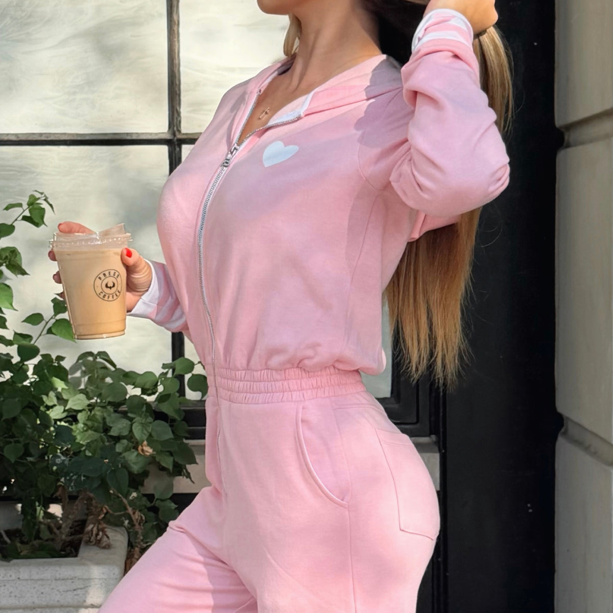 A woman wearing a light pink zip up tracksuit with a white heart on the chest, posing outdoors and holding a coffee cup.