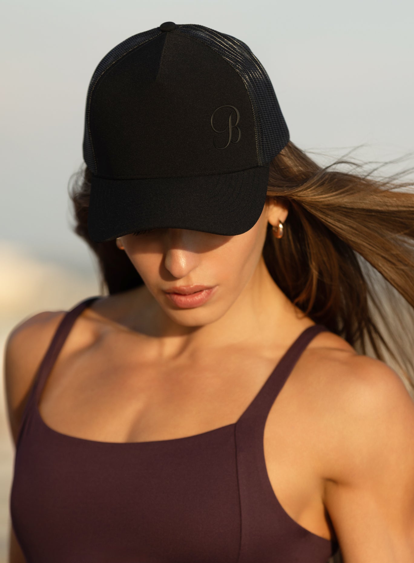 A woman wearing a black trucker-style baseball cap with a tonal embroidered monogram logo.