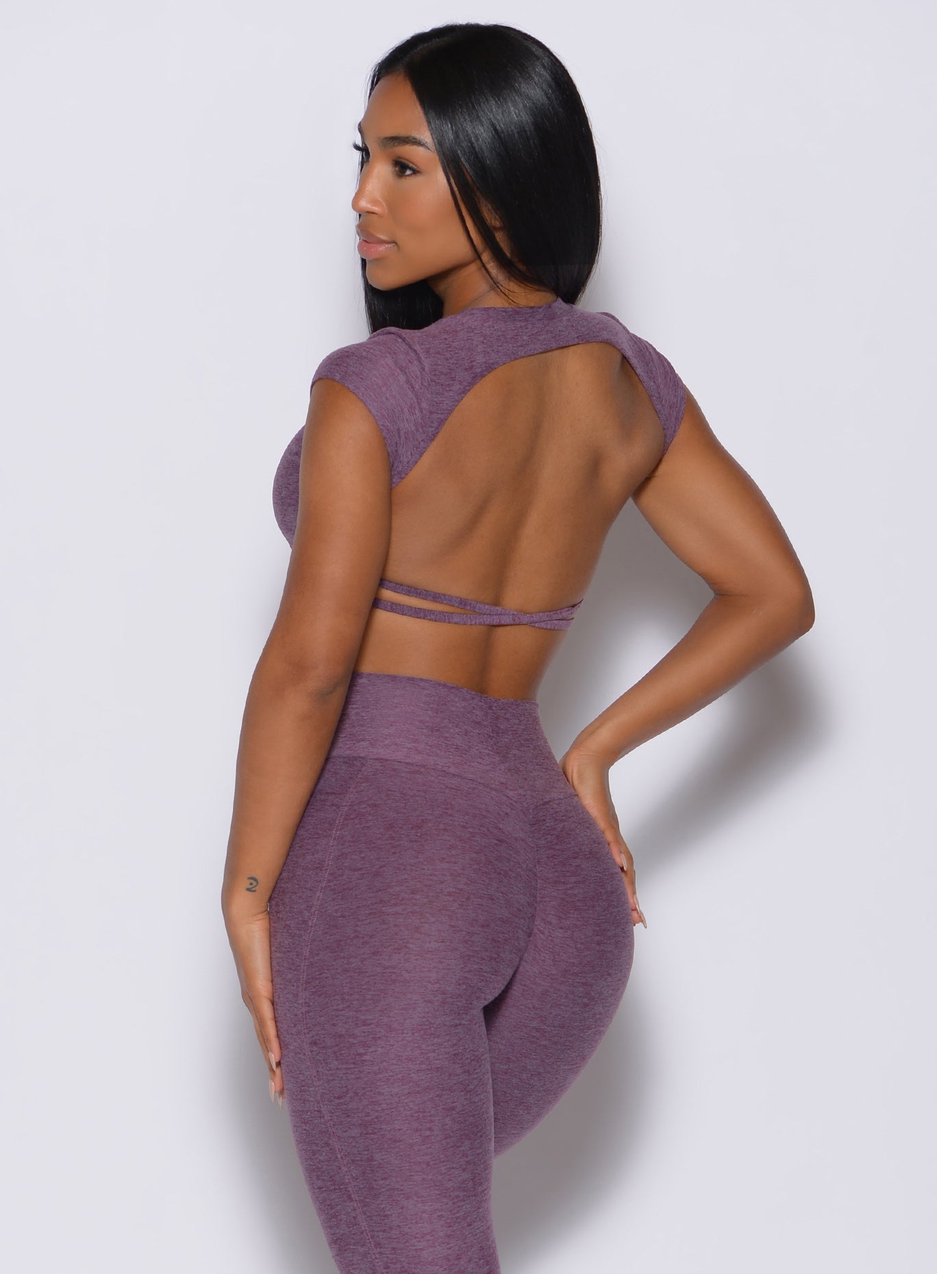 Back profile view of a model facing to her left wearing our open back tee in Regal Purple color and a matching leggings