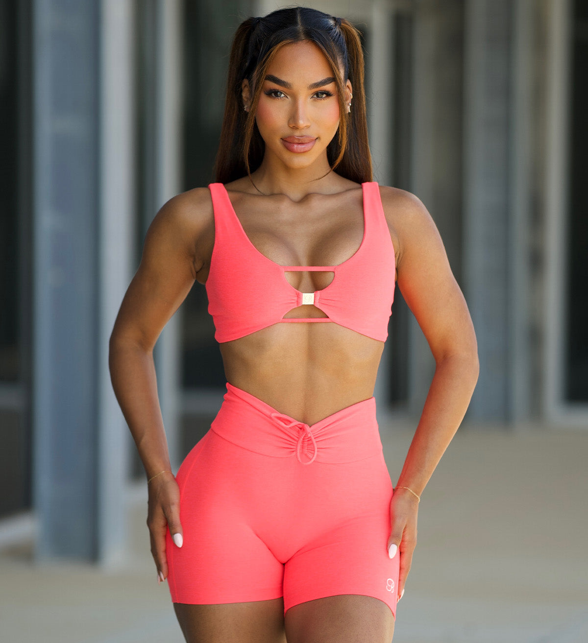 Woman with pigtails at the gym. She's wearing a neon pink sports bra with keyhole details and matching fitted athletic shorts.