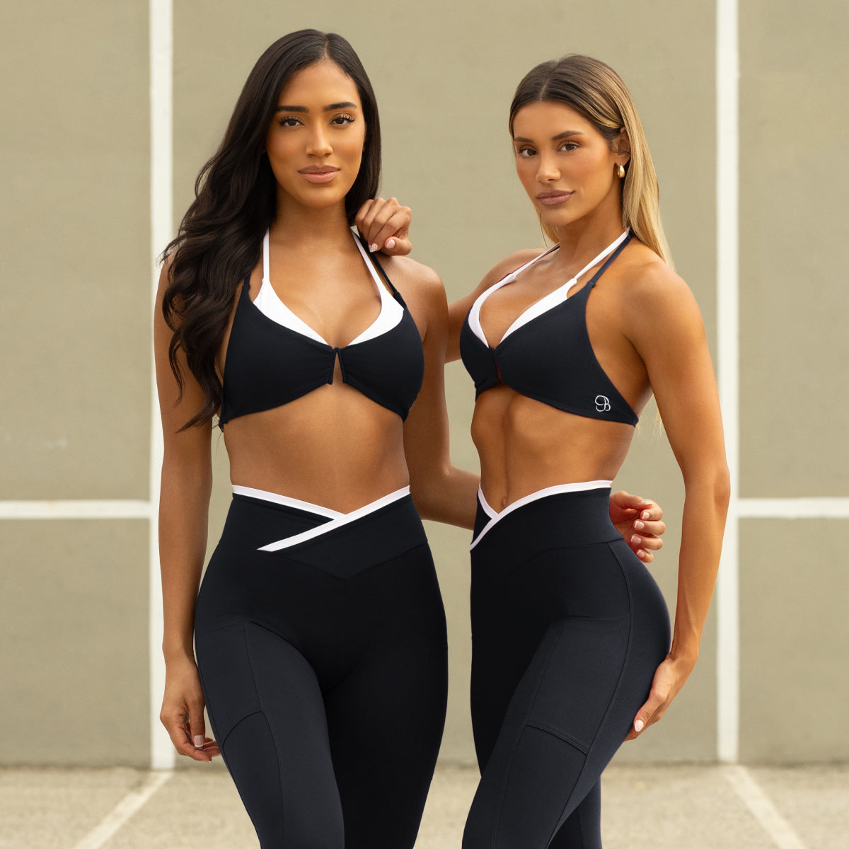 Two women wearing black sports bras and leggings with white accents on a neutral background.