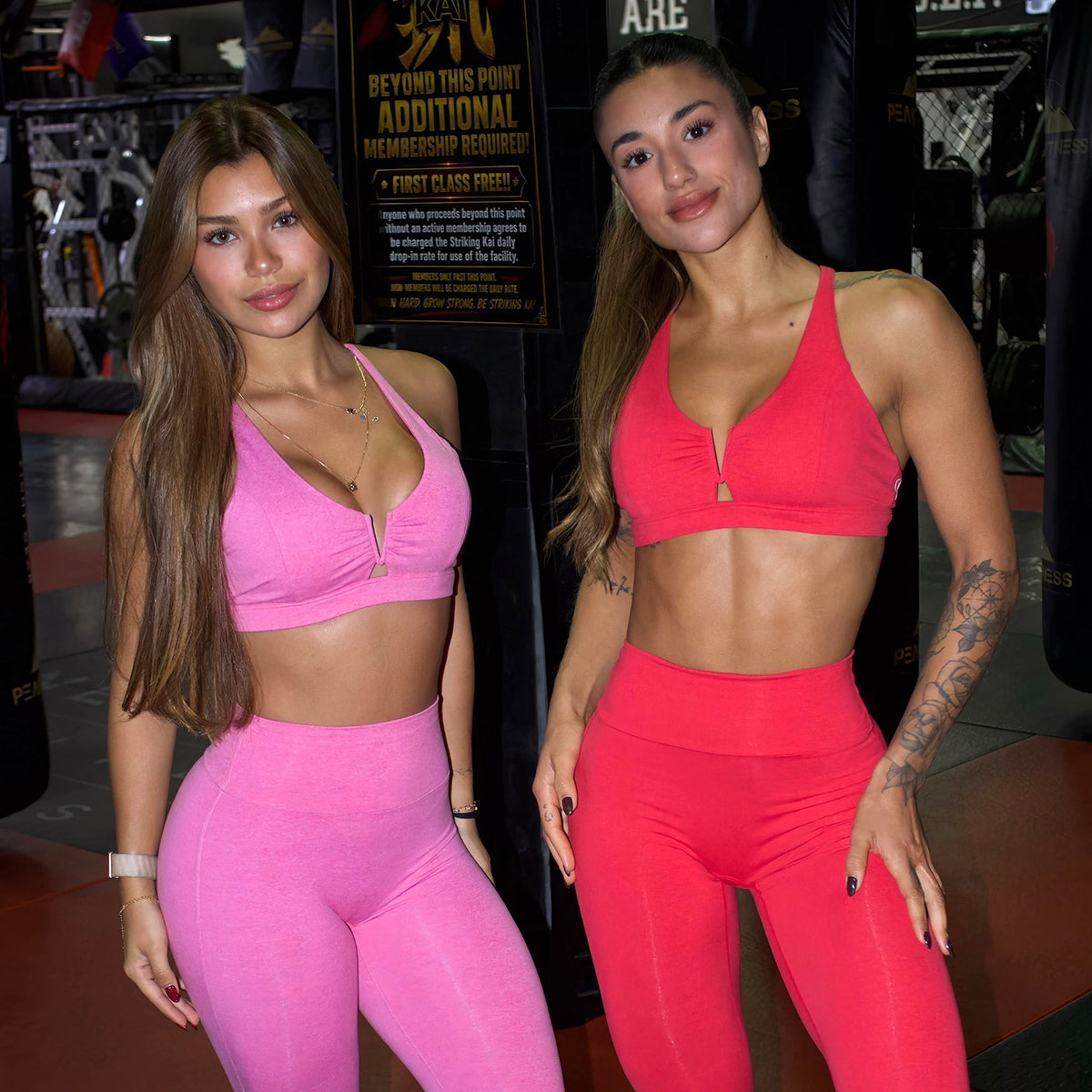 Two women posing in matching athletic wear at a gym. The woman on the left wears a bright pink sports bra and high-waisted leggings, while the woman on the right wears a coral-red sports bra and matching leggings.