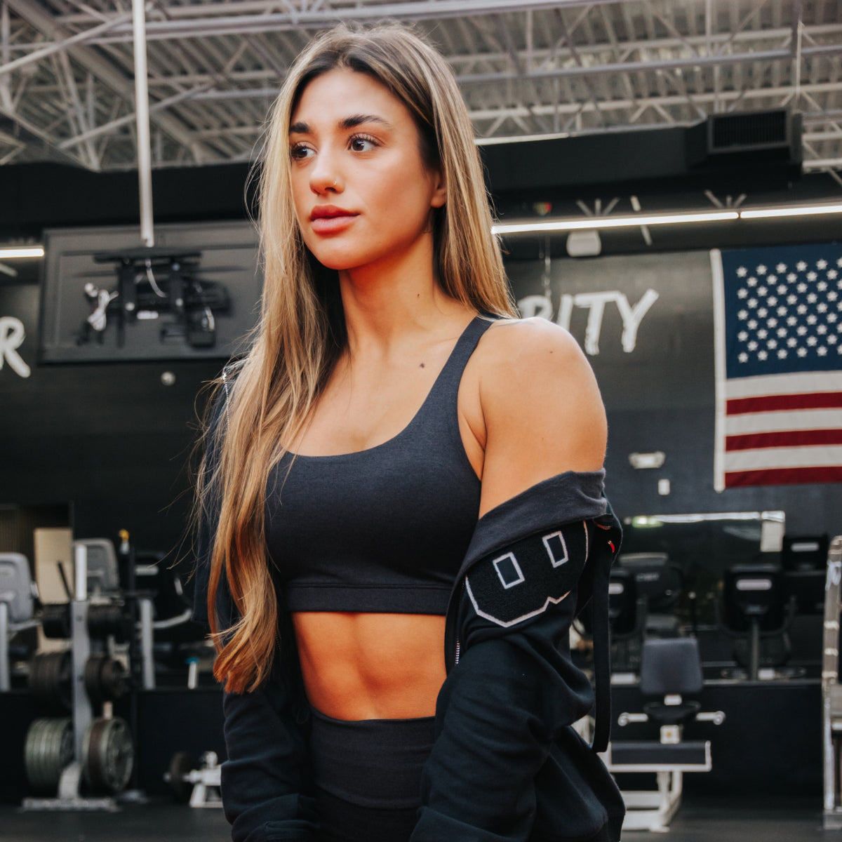 A woman in a gym wearing a dark gray sports bra and black athletic jacket, with a black sports bra and matching leggings visible. The background shows gym equipment and an American flag on the wall.