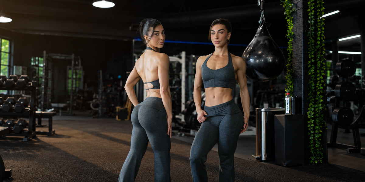Two women in matching gray athletic sets stand in a gym, one facing away to show the back of the sports bra and leggings and the other facing forward showing the front. A black hanging punching bag and gym equipment are visible in the background.