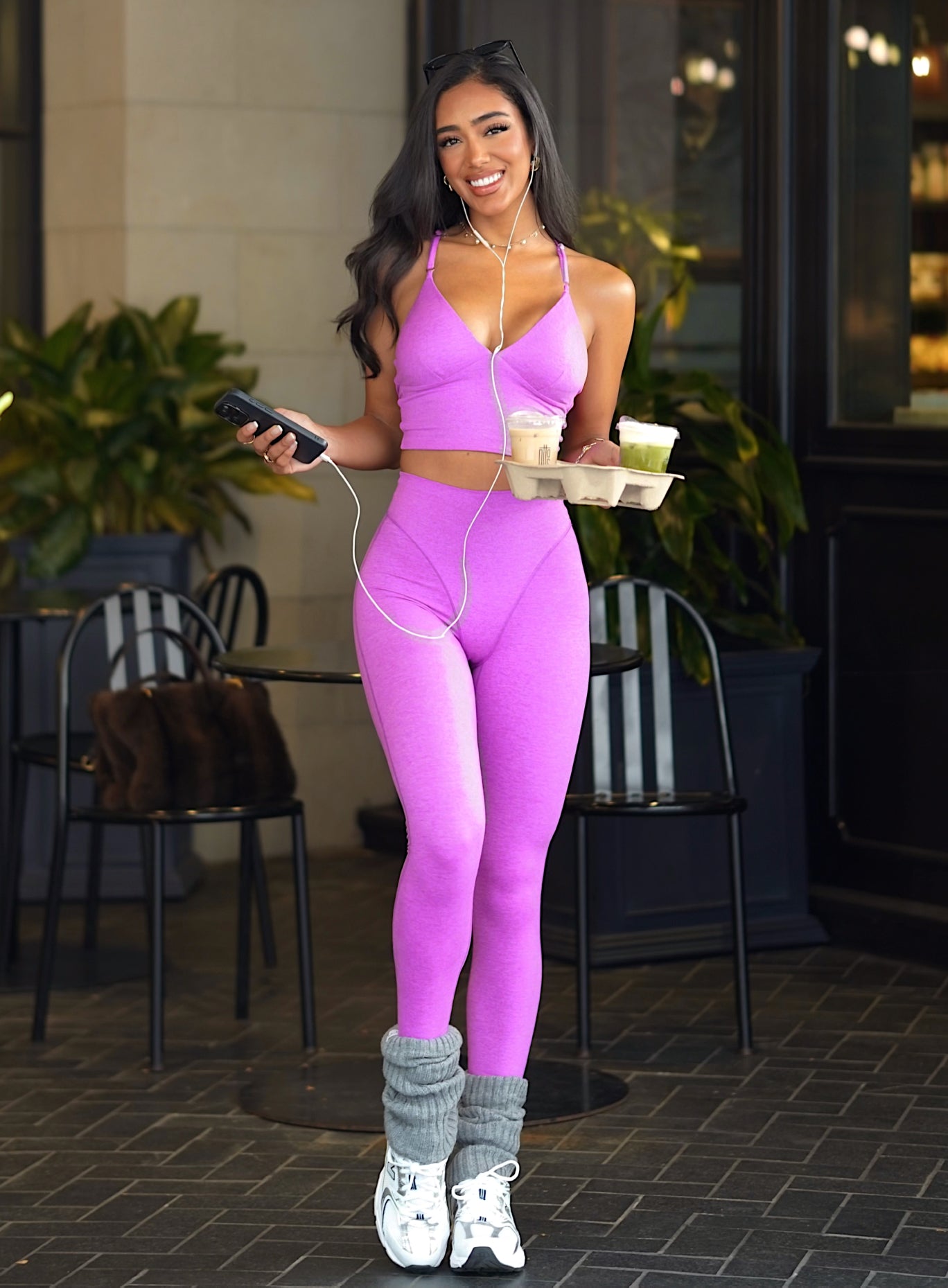 Woman in a pink outfit holding a tray with drinks outdoors wearing purple activewear bra and matching leggings with gray leg warmers and white sneakers