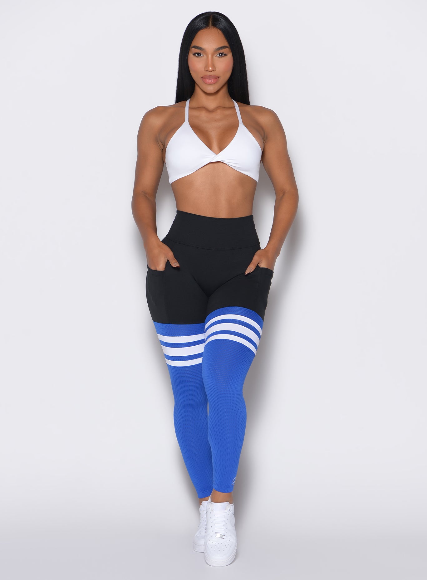 Front profile shot of a model with both hands in the pockets wearing the Perform Thigh Highs in Black Sky color along with a white sports bra