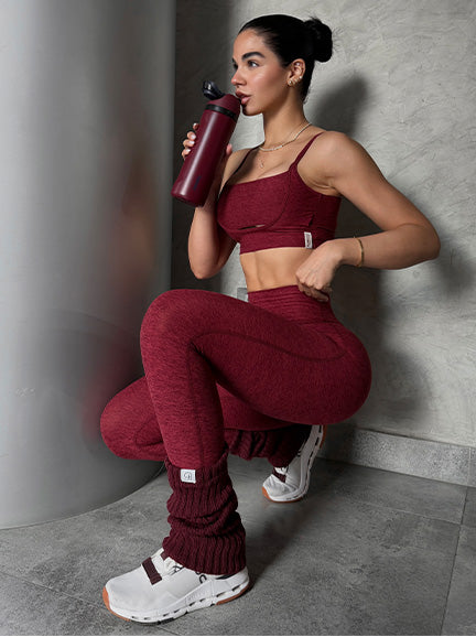 Woman in maroon athletic wear holding a water bottle against a gray wall.