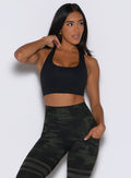 front profile view of a model with one hand inside the side pocket of the leggings wearing the Reversible Tank Bra in Green/Black camo