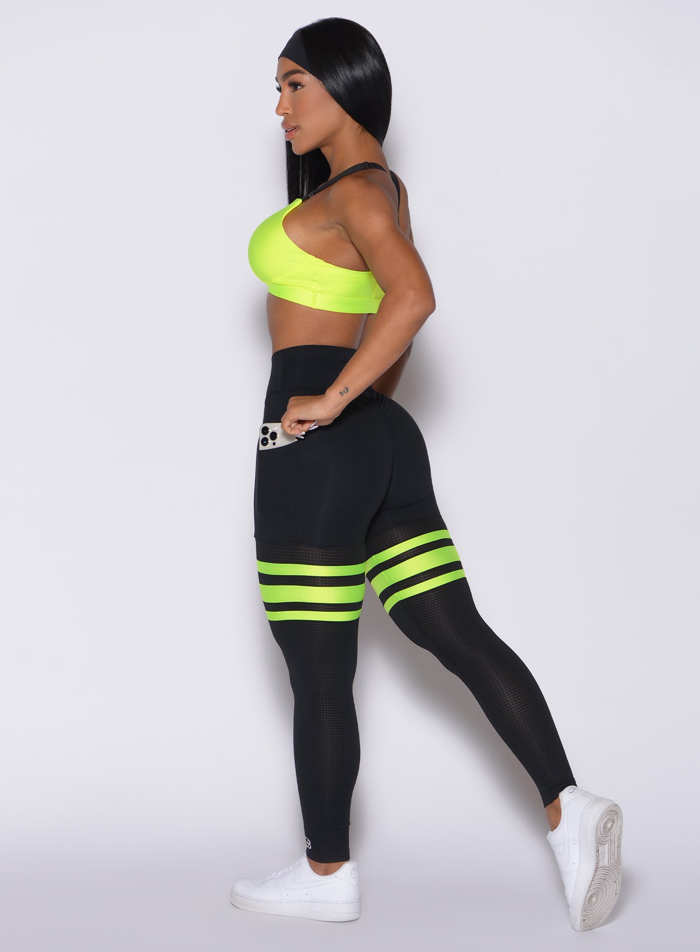Left side profile view of a model taking her phone out of her left pocket wearing our scrunch thigh high in black with neon yellow stripes on the thighs and a matching sports bra
