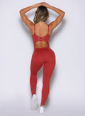 back side view of a model with both hands resting on top of her head wearing the Sculptura Bodysuit in Sunrise red color