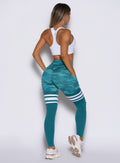 back side profile view of a model with both hands inside the side pockets wearing the Scrunch Thigh Highs in Teal camo color