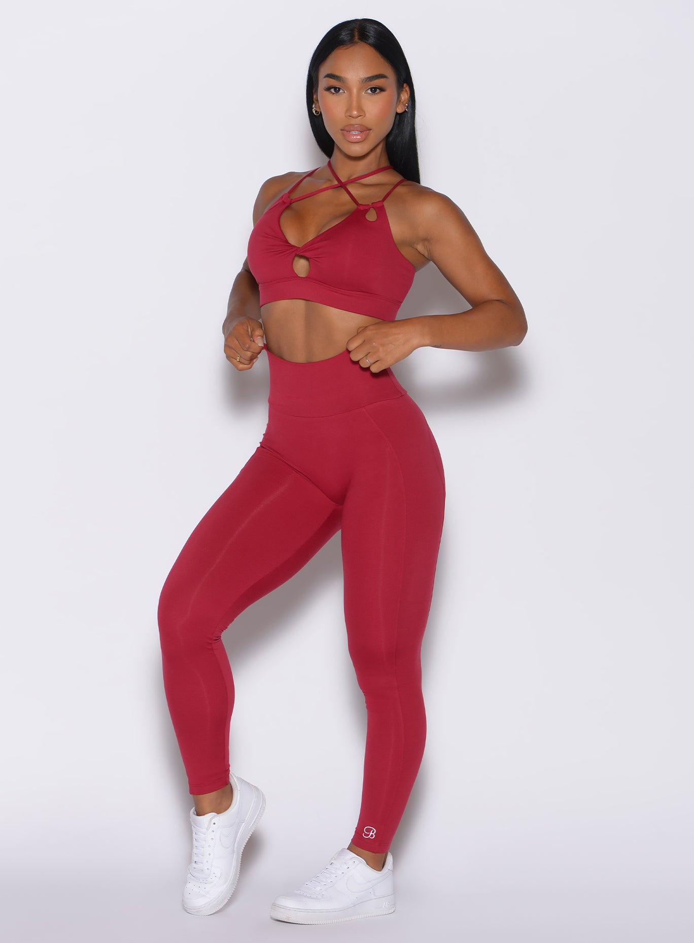 Front profile view of a model adjusting the waistband of the shape leggings in maroon color, that she is wearing,  along with the matching bra