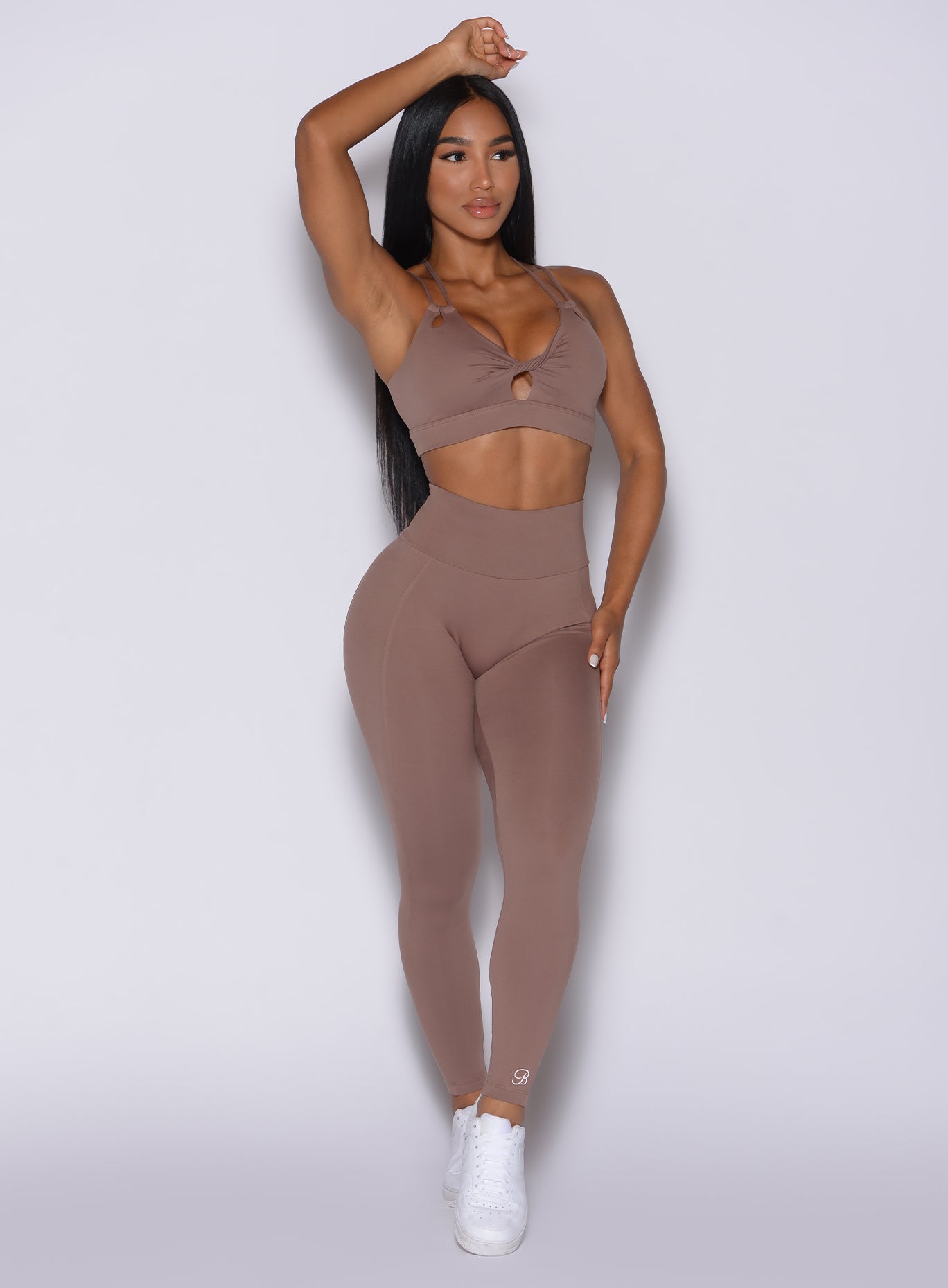 Front profile view of a model with her right hand over her head wearing our new and enhanced shape leggings in tan color and a matching bra