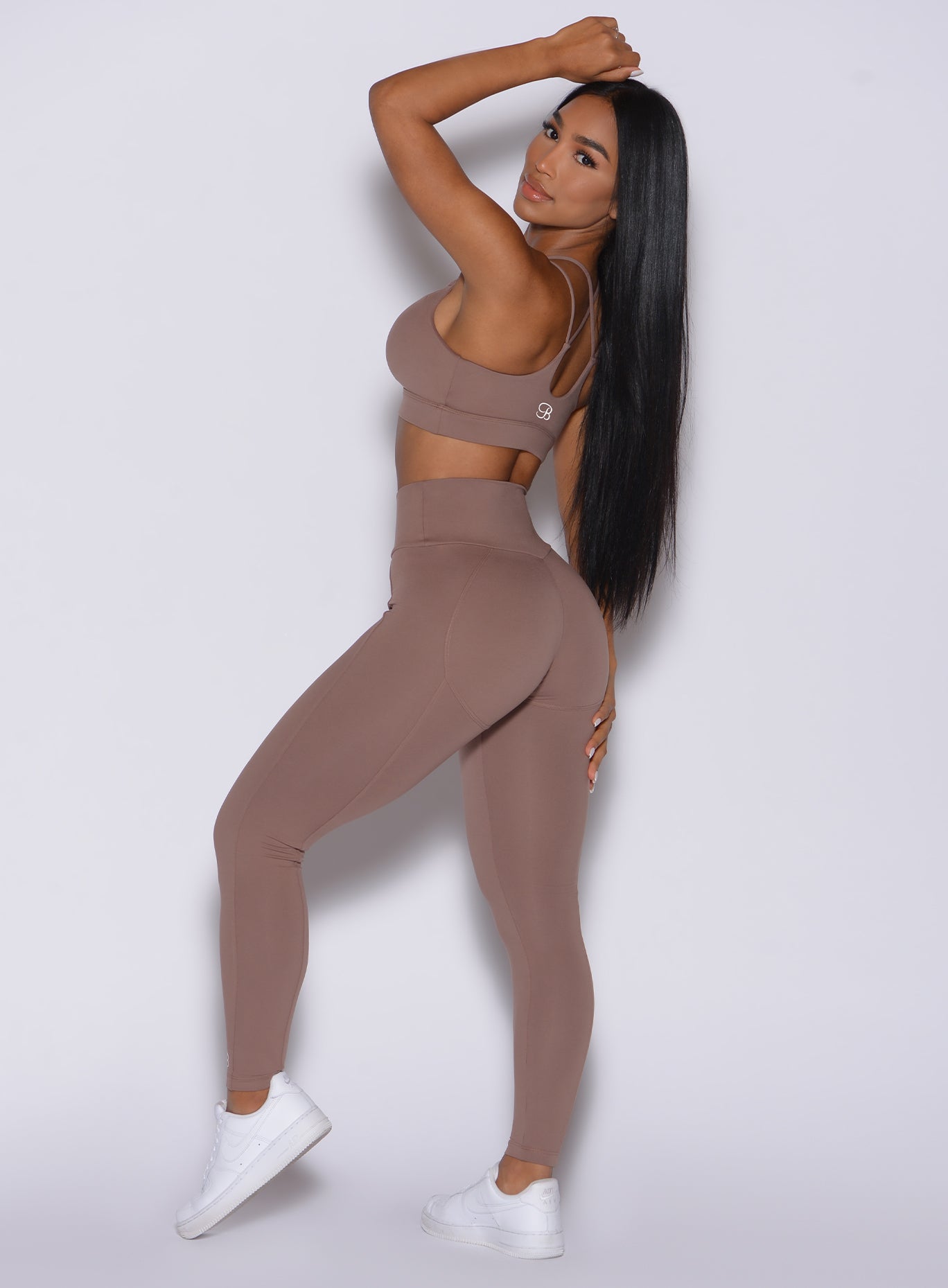 Left side profile view of a model facing to her left wearing our new and enhanced shape leggings in tan color and a matching sports bra