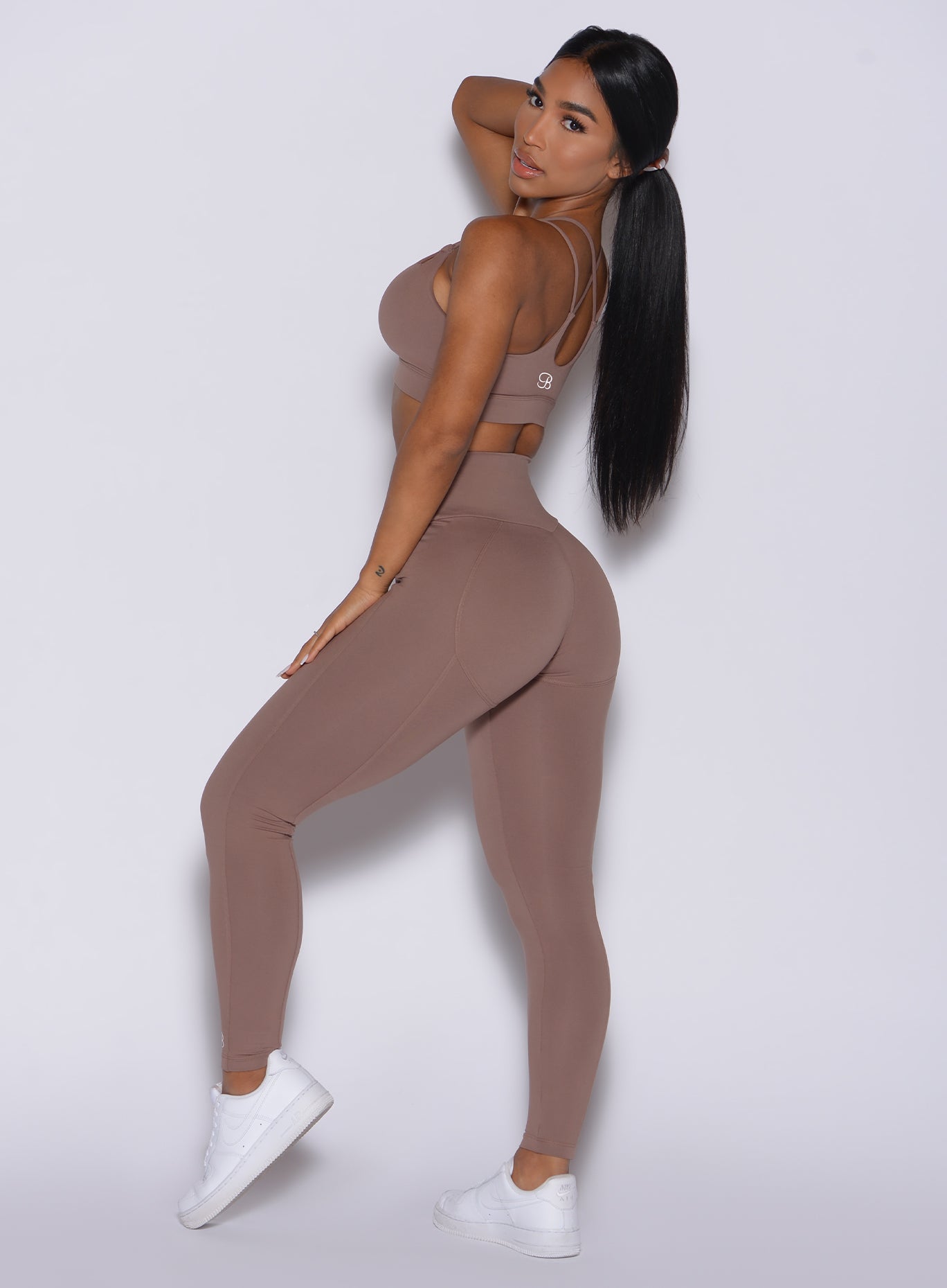 Left side profile view of a model holding her hair by her right hand wearing our new and enhanced shape leggings in tan color and a matching bra