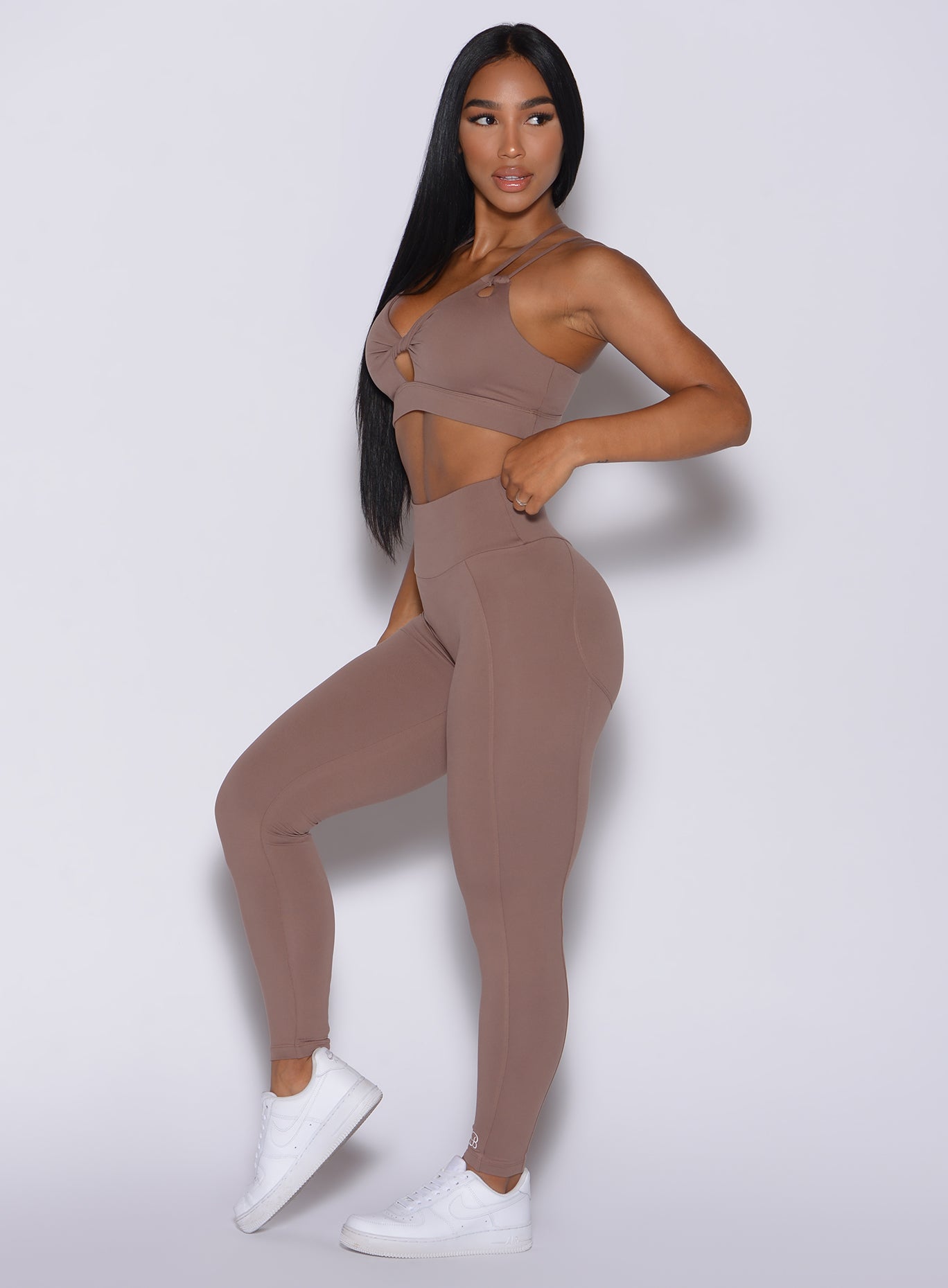 Left side profile view of a model angled slightly to her left wearing our new and enhanced shape leggings in tan color and a matching bra