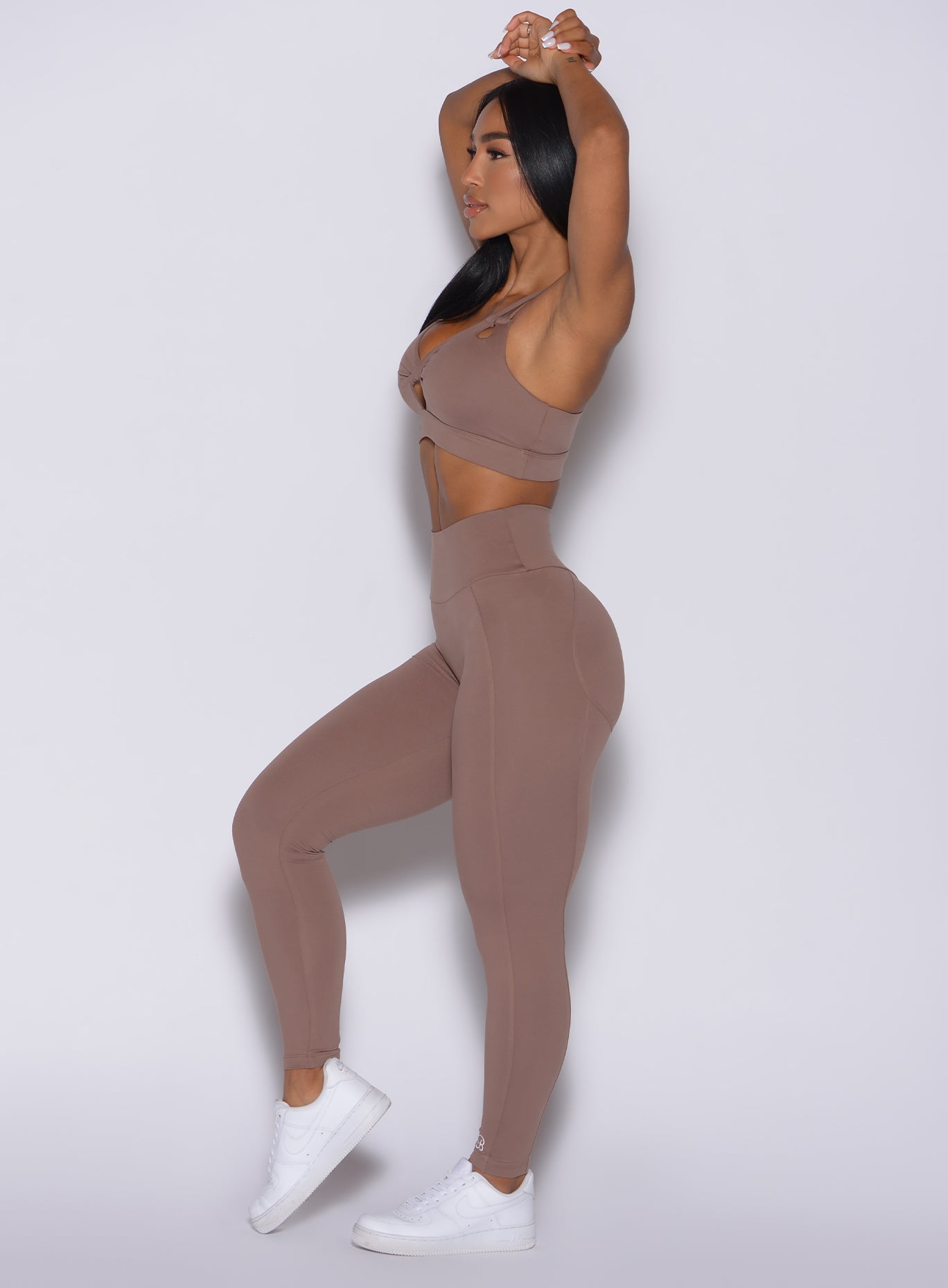 Left side profile view of a model with both her hands over her head wearing our new and enhanced shape leggings in tan color and a matching bra