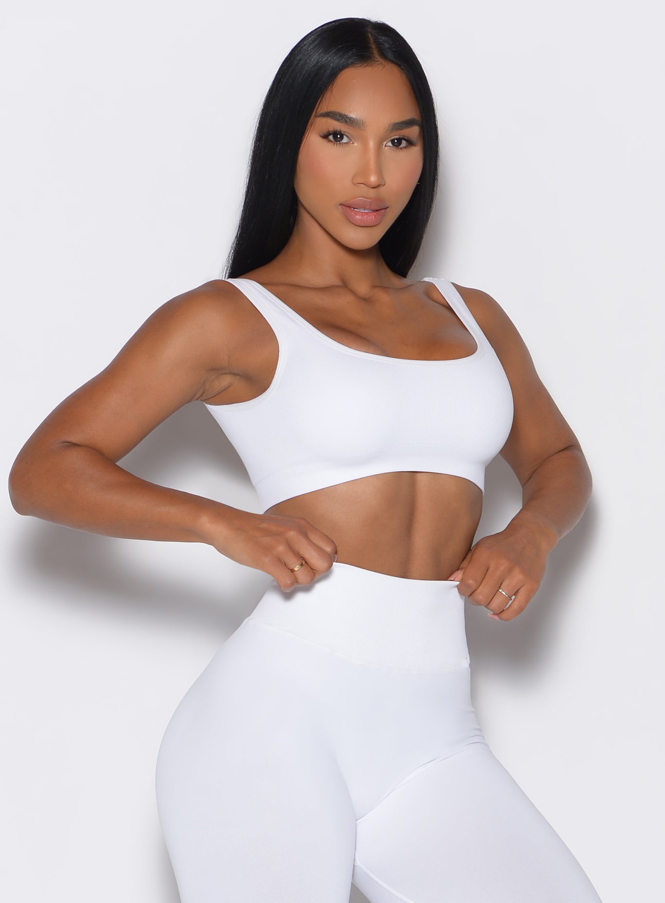 Front profile view of a model facing forward, adjusting the waistband on her leggings, and wearing our white square neck bra