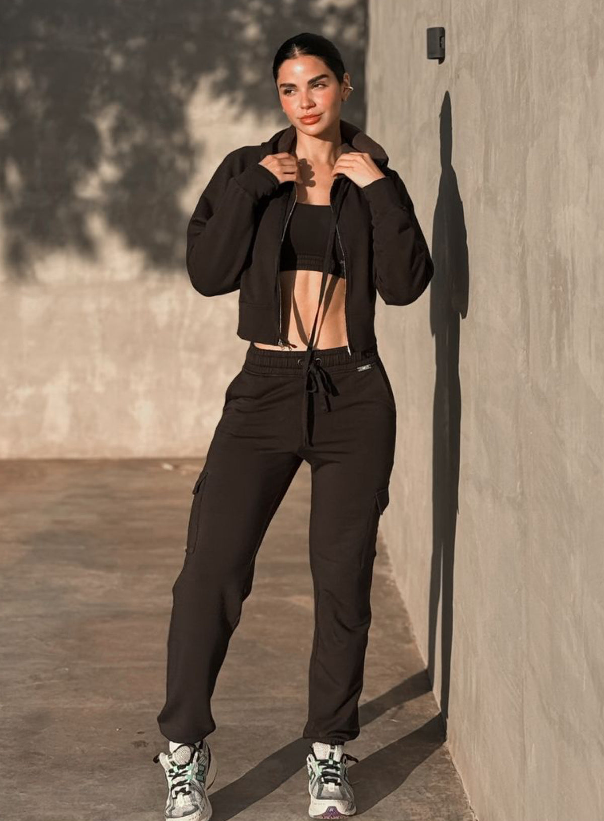 A woman stands outdoors against a textured concrete wall wearing a black cropped zip-up hoodie, black sports bra, black jogger pants, and white sneakers with green accents.