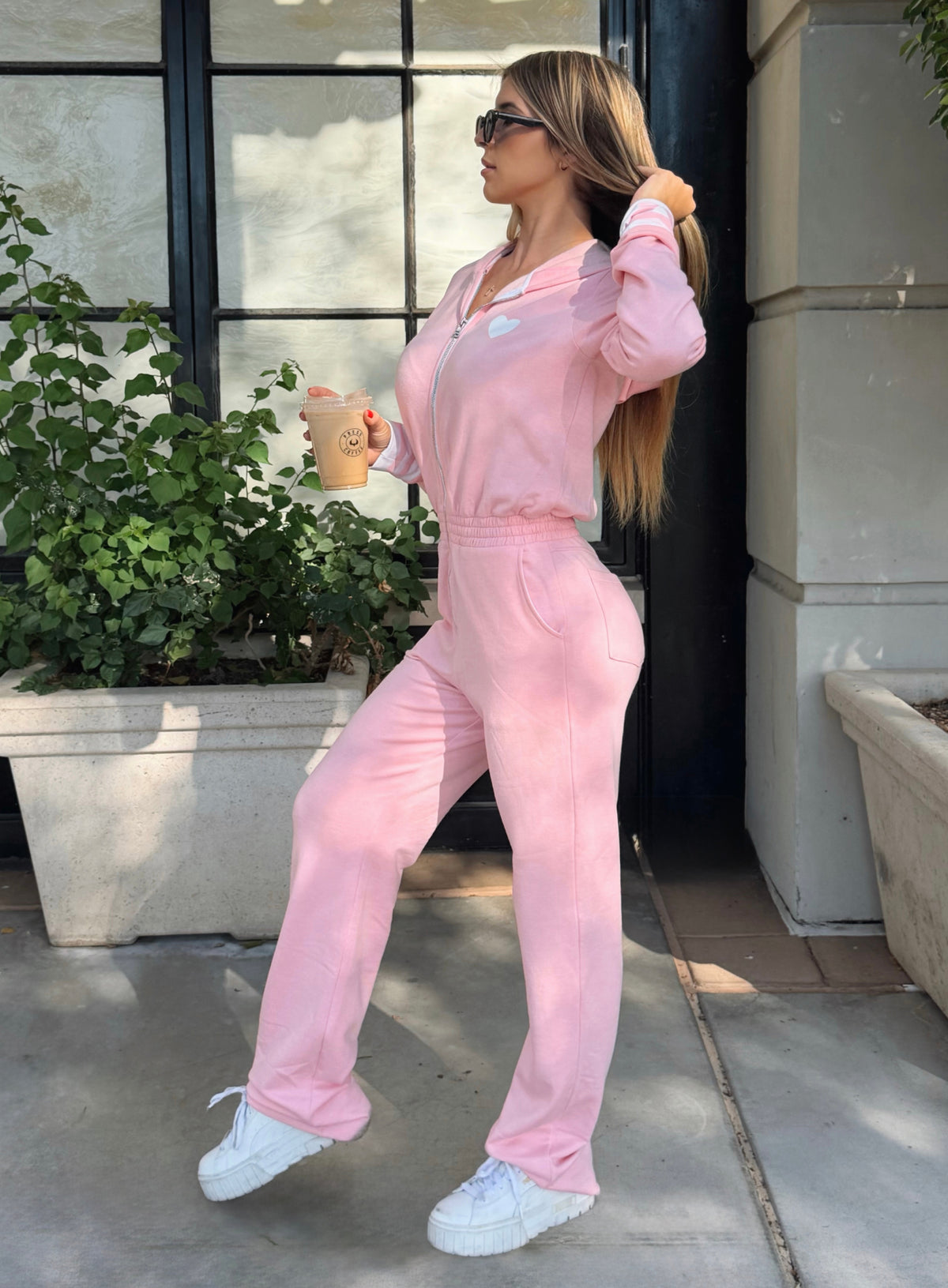 A woman wearing a light pink lounge fully body jumpsuit, styled with white sneakers and sunglasses while holding a coffee cup outdoors.
