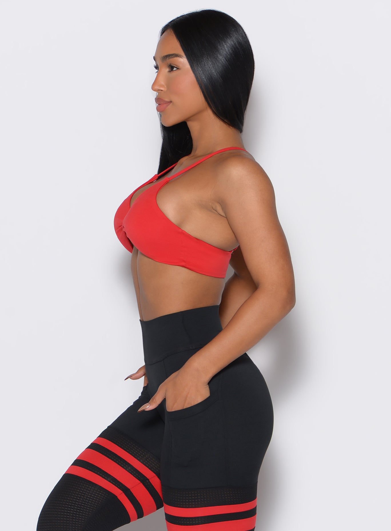 Left side profile view of a model with both her hands in the pockets of her leggings wearing our Twist Mini Bra in Fire color