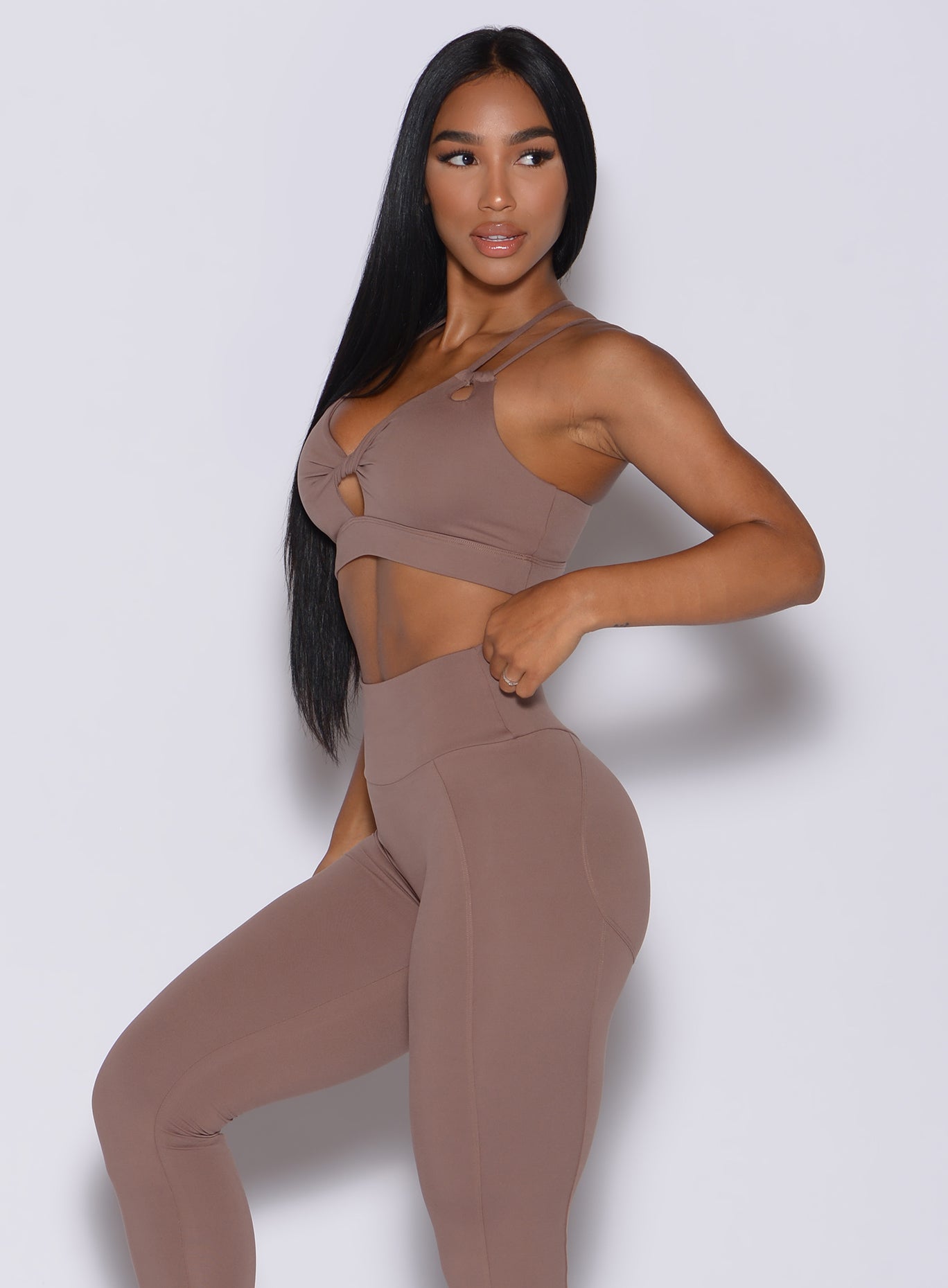 Left side profile view of a model angled left wearing our twist sports bra in tan color and a matching leggings